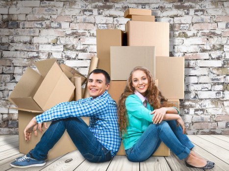 A young man and woman sit on a light wooden floor in front of a weathered exposed brick wall, surrounded by several cardboard boxes of various sizes, some closed, others partially open, suggesting a recent move or packing activity. The man, positioned on the left, is wearing a blue and white checkered shirt and dark jeans, with short dark hair, and is smiling while leaning back slightly with his arm resting on his knee. The woman, on the right, has long, curly, light brown hair and is dressed in a light teal sweater and blue jeans, also smiling as she crosses her arms and sits with her legs stretched out. Behind them, the boxes are stacked with some leaning, indicating an active packing or unpacking process typical of house removals in South Kensington. The environment appears to be an interior space, possibly a living room or hallway, with natural light illuminating the scene. This image, associated with South Kensington Man with Van, reflects a professional, local removals service engaged in household moves within the area, highlighting the careful handling and organization involved in domestic relocation tasks.