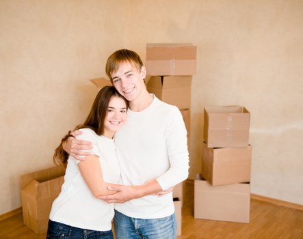 A young man and woman with light skin, both dressed in white tops, are standing close together indoors in front of a plain beige wall. They are smiling and embracing each other, with the woman slightly in front of the man. Behind them, several cardboard moving boxes of various sizes are stacked against the wall, some sealed with packing tape. The boxes appear to be filled with household belongings and are arranged on a wooden floor, indicating a residential setting. The environment suggests that they are in the process of packing or preparing for a house move, which aligns with house removals services in South Kensington. The lighting is natural and even, highlighting the clarity of the scene, capturing the moments before or after relocation activities.