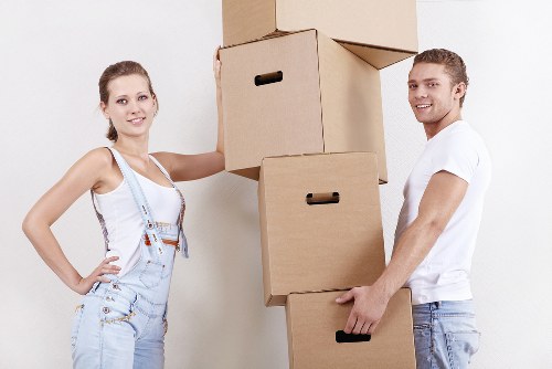 A young woman and a young man are indoors, standing next to a stack of four cardboard boxes of various sizes, which they are holding and supporting together. The woman, on the left, has light brown hair tied back and is wearing a white sleeveless top and light blue overalls with a visible measuring tape hanging from her neck. The man, on the right, has short brown hair and is dressed in a white t-shirt and light denim jeans, gripping one of the boxes with both hands. The background features a plain, neutral-colored wall, suggesting an interior space, possibly a residential setting in South Kensington, suitable for house removals. The boxes appear to be empty or packed, with some flaps open, ready for moving purposes. The lighting is bright and evenly distributed, highlighting the casual attire of the individuals and the simple, unmarked corrugated cardboard boxes. This scene captures a moment of preparation for a house move, typical of services provided by South Kensington Man with Van, emphasizing professionalism and careful handling of household items during relocations in the local area.
