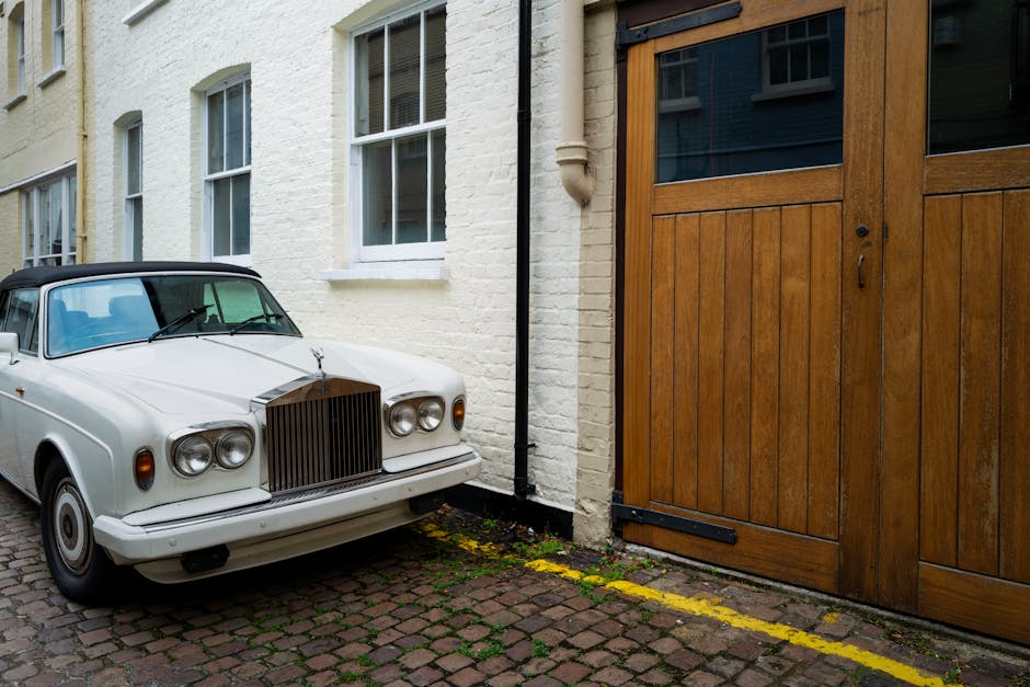 A white vintage car with a black soft top is parked on a narrow cobblestone driveway beside a white brick house with double-pane sash windows. The vehicle is positioned close to a large wooden sliding