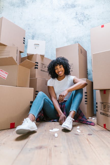 A man with curly hair and medium skin tone, sitting on a wooden floor in a brightly lit room with large arched windows, surrounded by multiple cardboard boxes sealed with red tape and bubble wrap. The