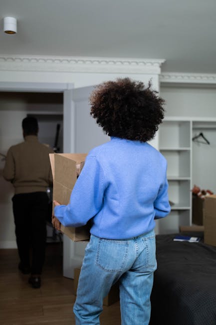 A young woman with long, curly brown hair is inside a room, seen lifting a large cardboard moving box with both hands, while partially leaning on a wooden chair behind her. She is wearing a light grey